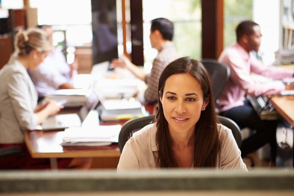 Businesswoman,Working,At,Desk,With,Meeting,In,Background